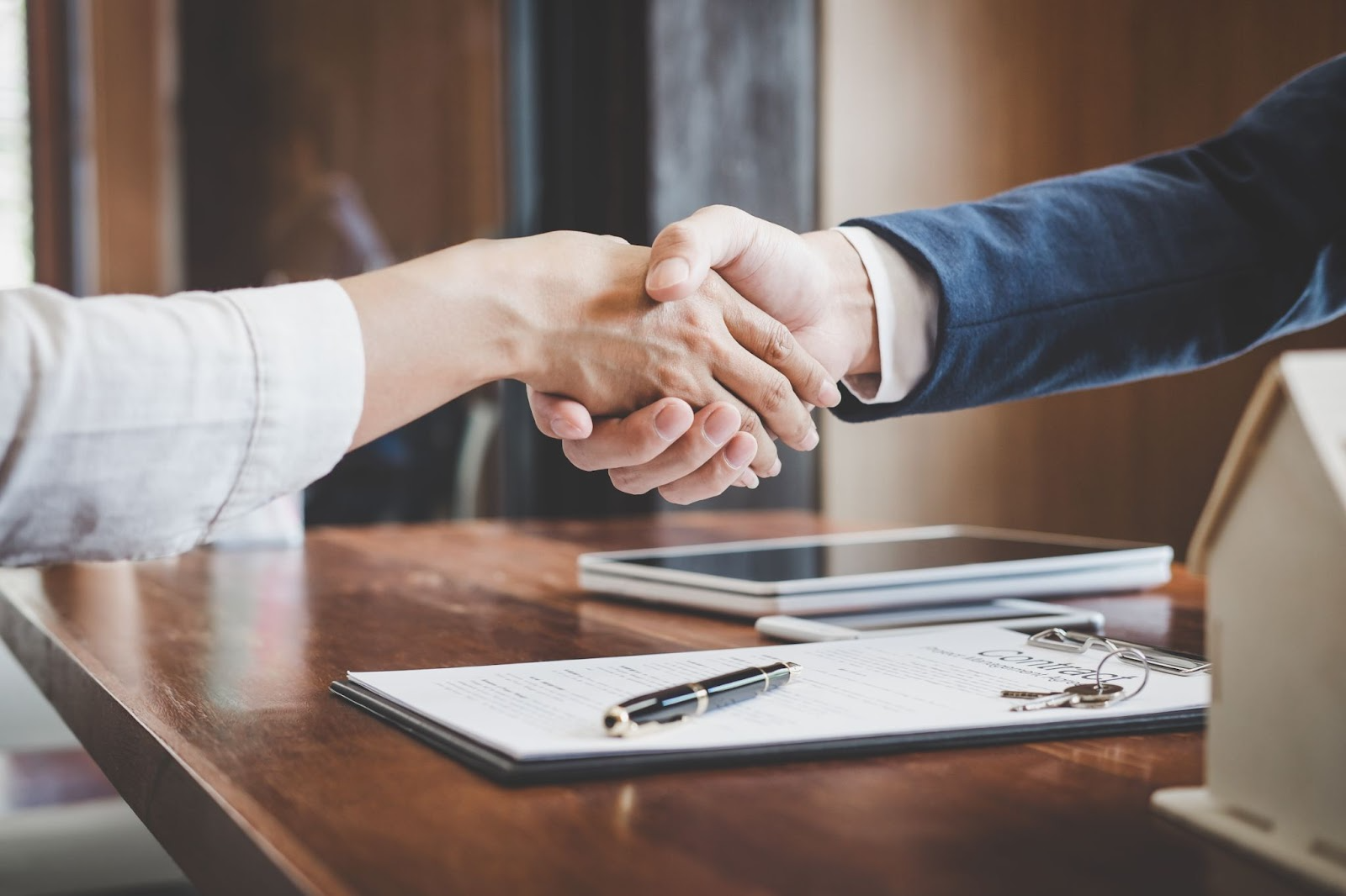Two people shaking hands over a contract with a pen and tablet on the table